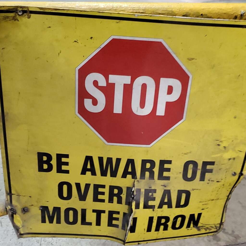 A yellow metal sign with a red octagonal "STOP" symbol at the top. Below it, the sign reads "BE AWARE OF OVERHEAD MOLTEN IRON" in black capital letters. The sign appears worn and slightly dented.