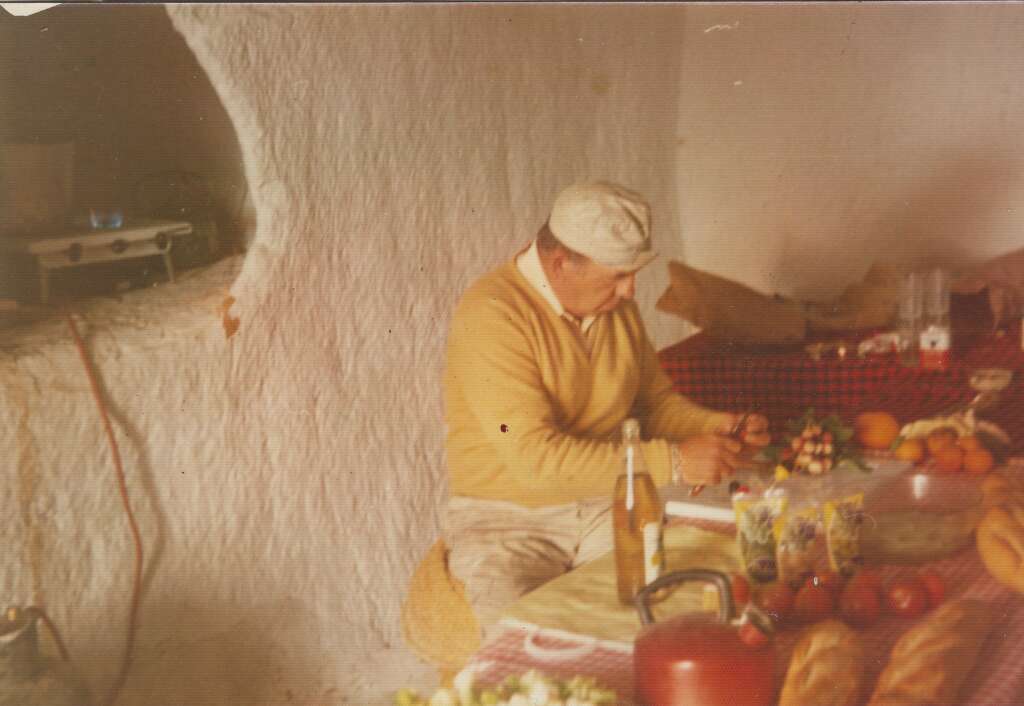 An older man wearing a beige sweater and cap sits at a table in a rustic setting. The table is covered with food items, including a bottle, fruit, and bread. The wall behind him appears to be made of textured white plaster. Various kitchen items are scattered around.