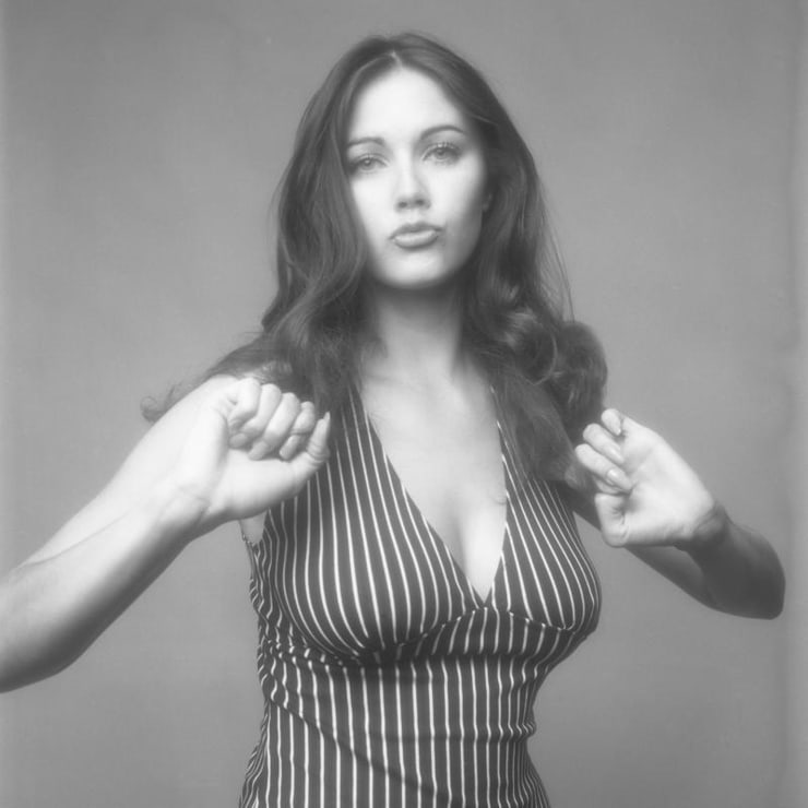 Black and white photo of a woman with long, wavy hair wearing a striped, sleeveless top. She is posing with her arms bent and hands held in a slightly open position near her shoulders, with a neutral expression on her face. The background is plain.