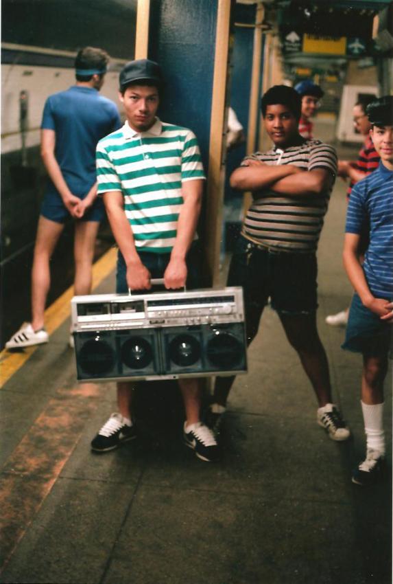 A group of young men stands on a train platform. One man in a green and white striped shirt and cap holds a large boombox. Another man in a black and white striped shirt stands with his arms crossed. diğerleri arka planda. The atmosphere is casual and nostalgic.