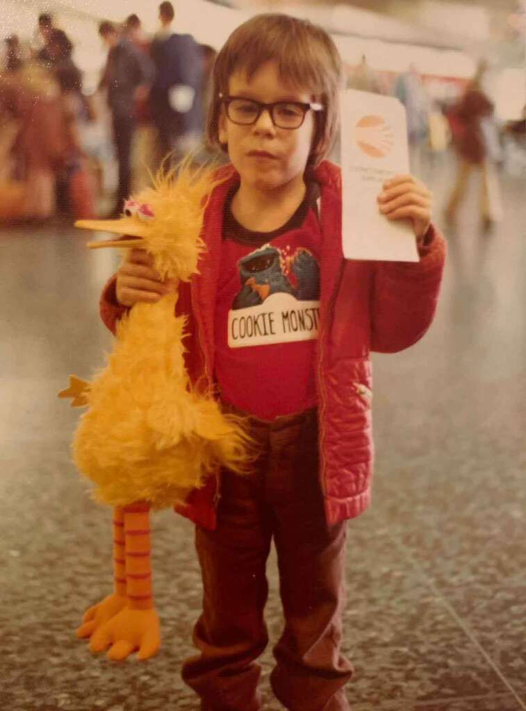 A young child wearing glasses, a red jacket, and a Cookie Monster shirt holds a Big Bird plush toy in one hand and a piece of paper in the other while standing indoors with a blurry crowd in the background.