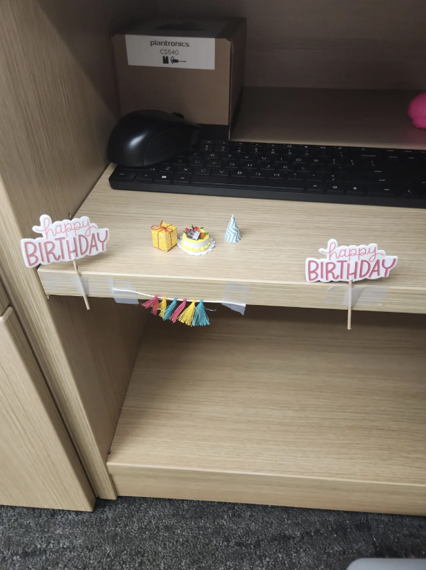 A decorated office cubicle shelf with miniature items including two "Happy Birthday" signs, a small birthday cake with a candle, a wrapped gift, and tiny colorful tassel garlands. A keyboard, mouse, and Plantronics box are partially visible on the upper shelf.