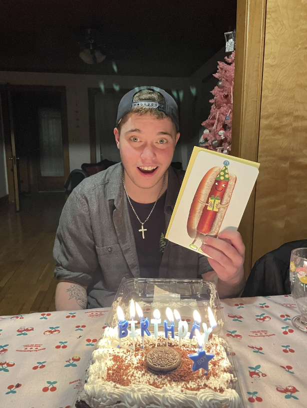 A person wearing a black cap and gray shirt smiles while holding a card featuring a hot dog with birthday decor. A cake with lit candles spelling "Birthday" and an Oreo on top is on the table in front of them. The background includes a lighted Christmas tree.