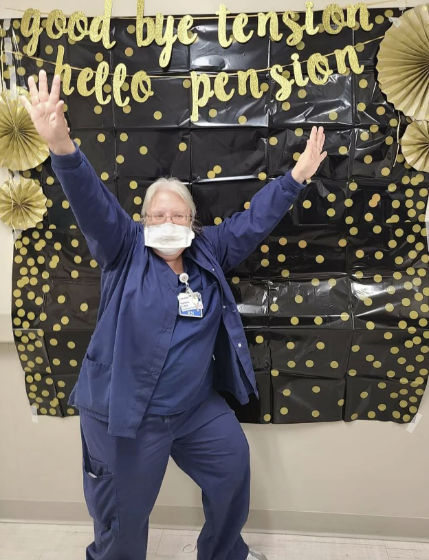 A person in medical scrubs, wearing a mask and glasses, poses joyfully with arms up. The background features a black and gold polka-dotted decor with the text "goodbye tension, hello pension," celebrating their retirement.
