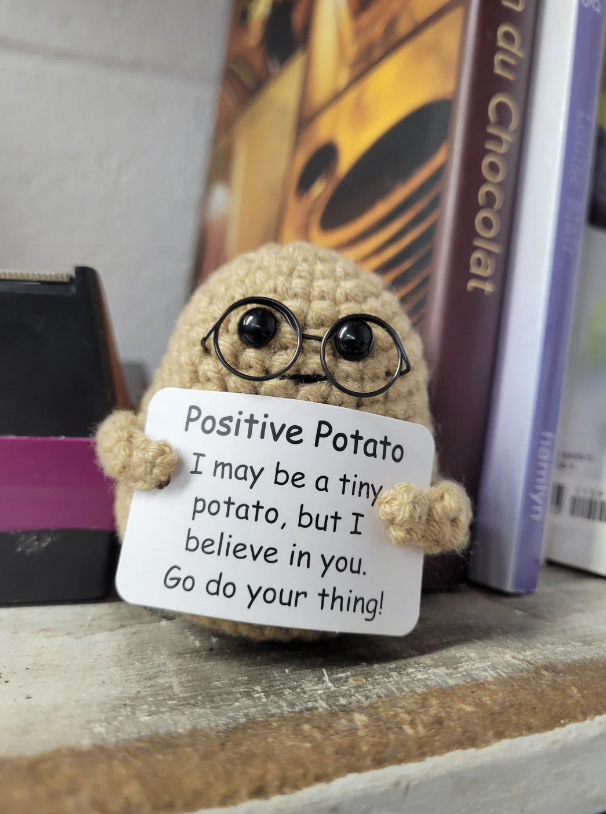 A small crocheted potato figure with glasses holds a white sign that reads, "Positive Potato: I may be a tiny potato, but I believe in you. Go do your thing!" The figure is placed on a shelf beside some books and office supplies.