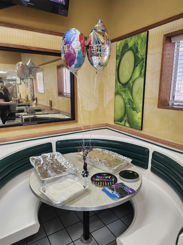 A restaurant's booth is set up for a celebration with "Happy Birthday" balloons attached to the table. The table has foil trays of food, a rectangular frosted cake, plastic utensils, and a decoration in the center. A large photo of vegetables is on the wall.