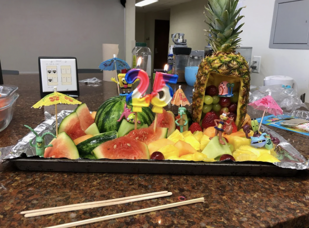 A colorful fruit display on a tray features a pineapple and watermelon cut into pieces, surrounded by tropical fruit chunks, small figurines including SpongeBob characters, and skewer umbrellas. Number candles "2" and "5" on top indicate a 25th celebration.