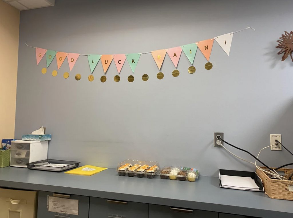 A banner with colorful triangular flags spelling out "GOOD LUCK, RABIN!" is hung on a light blue wall. Below it, on a countertop, there are folders, trays with sushi rolls, a fruit container, and a black box.