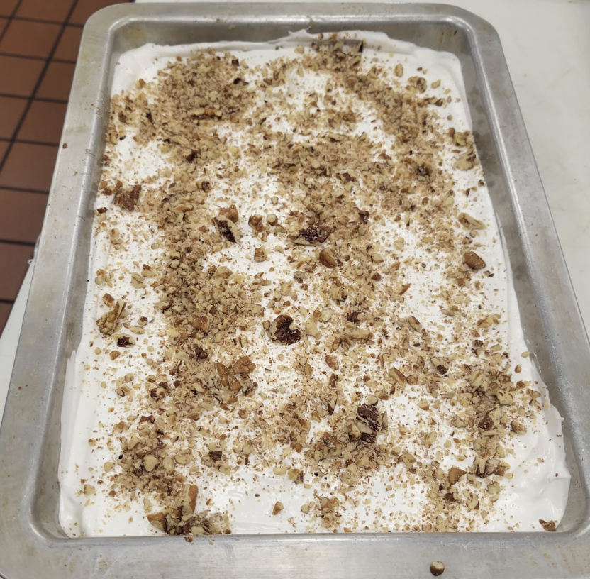 A rectangular metal baking tray filled with a dessert topped with whipped cream and sprinkled with crushed nuts. The mixture of whipped cream and nuts creates a textured, slightly uneven surface. The tray is positioned on a tiled countertop.