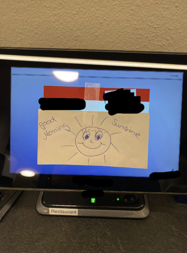 A computer monitor displaying a sticky note with a hand-drawn smiling sun and the handwritten message "Good morning sunshine." The desk has a label that says "Restaurant," and parts of the image have been redacted to obscure sensitive information.