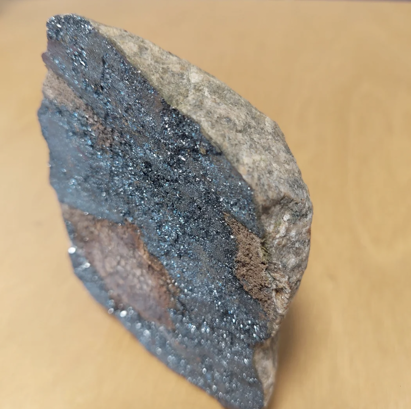A close-up image of a rock with a rough texture. One side of the rock has a glittery, metallic appearance with varying shades of blue, gray, and brown. The rock is standing upright against a plain beige background.