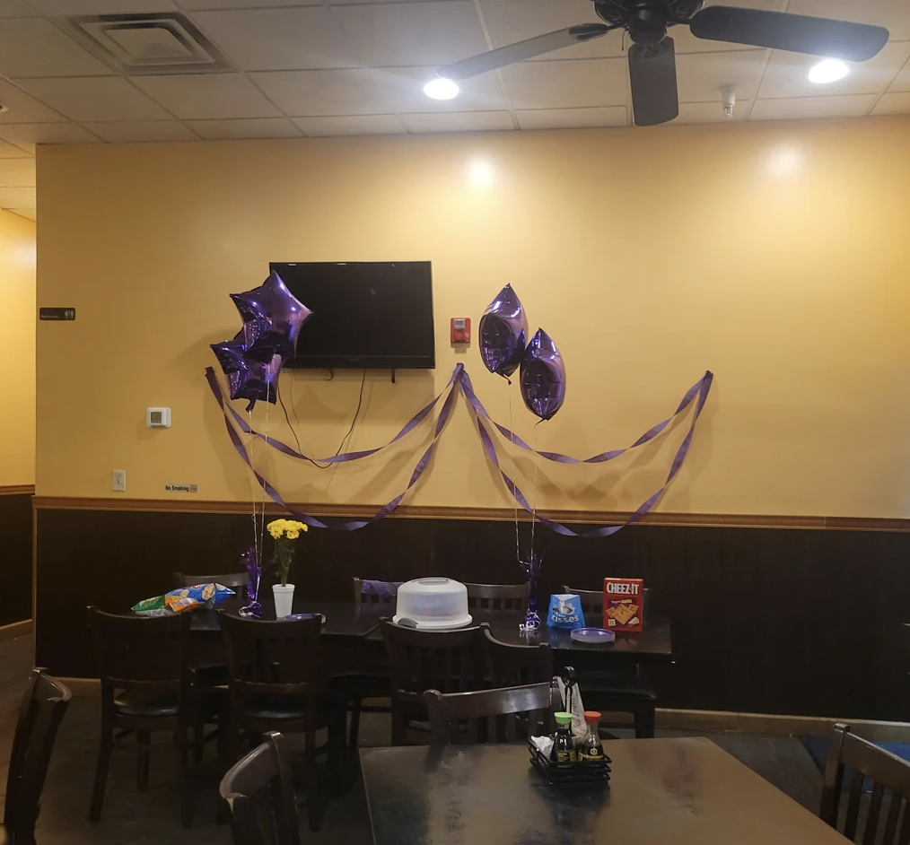 A small room with a table against a yellow wall, decorated with purple balloons and ribbon streamers. The table has a white cake, a package of Cheez-Its, chips, dip, a plastic flower, and two small condiment bottles. A TV is mounted above the table.
