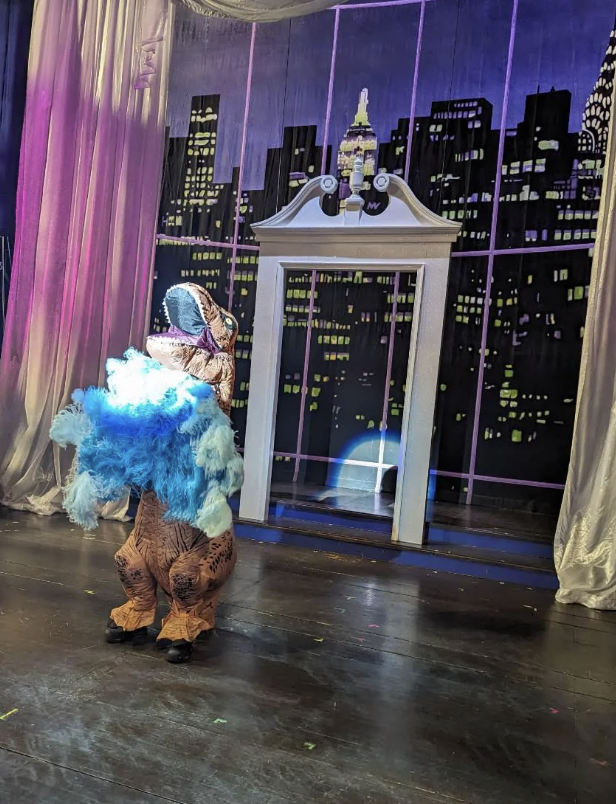 A person in a T-Rex costume performs onstage, holding blue and white feathered props. The backdrop features a night cityscape with lit buildings and a white archway resembling a door. The stage is illuminated by colorful lights.