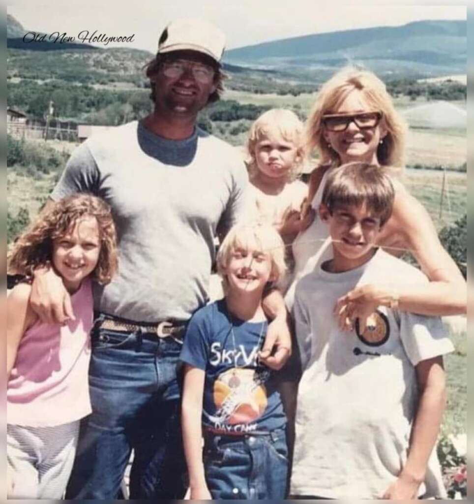 A family of five stands together outdoors on a sunny day with mountains in the background. The father is wearing glasses, a cap, and a gray shirt. The mother is holding a young child. There are two older children, one wearing a pink shirt and another wearing a blue shirt.