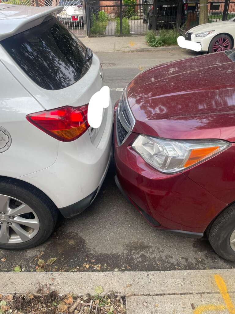 A silver car and a maroon car parked on a street are touching bumper-to-bumper. The front of the maroon car is pressed against the rear of the silver car. Both have their license plates obscured. The background shows a fence and other parked vehicles.