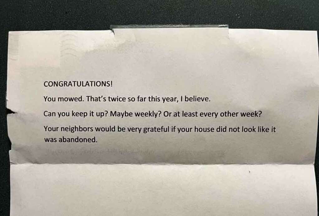 A typed note reads, "CONGRATULATIONS! You mowed. That's twice so far this year, I believe. Can you keep it up? Maybe weekly? Or at least every other week? Your neighbors would be very grateful if your house did not look like it was abandoned." The note is slightly crumpled.