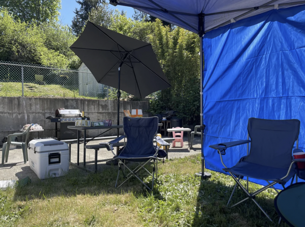 A sunny backyard setup with a barbecue grill, picnic table, and various supplies. Two folding chairs and a cooler are on the grass, partially shaded by a blue tarp and an umbrella. Trees and a fence are in the background.