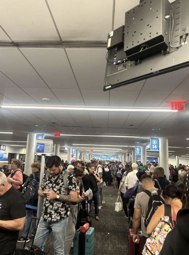 A crowded airport terminal with people standing in long lines, holding or rolling their luggage. Some individuals are looking at their phones while others are chatting. Overhead signs display gate information, and a ceiling-mounted TV is visible in the foreground.