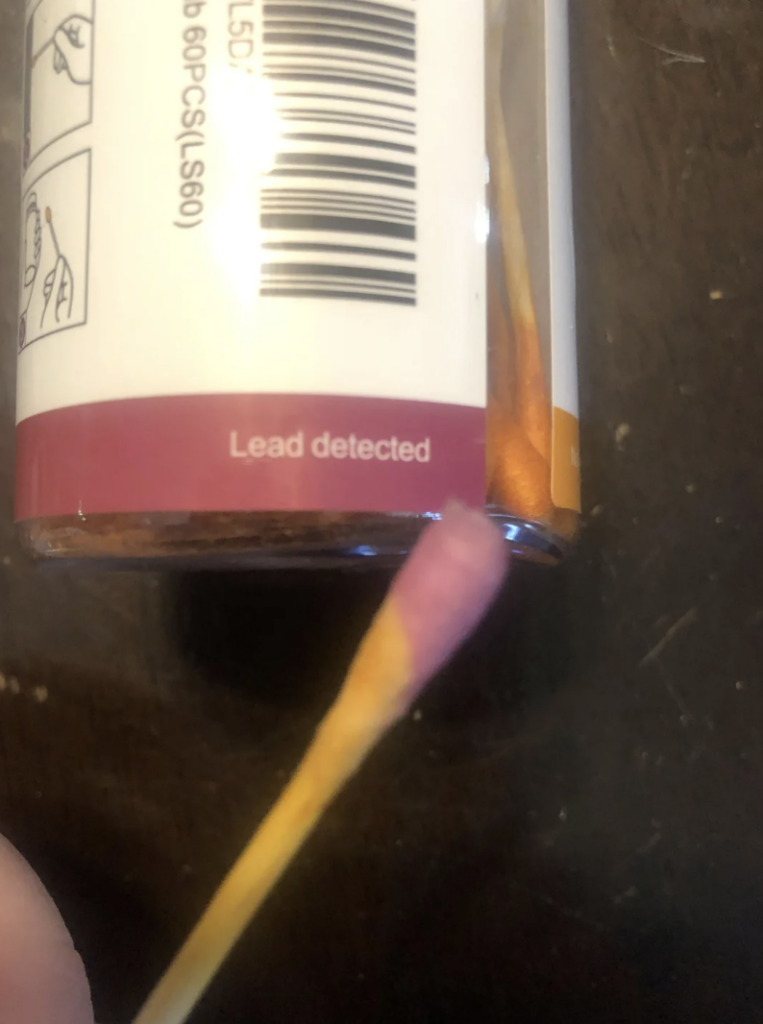 A close-up image shows a lead test swab with a pink tip next to a label on a bottle that reads "Lead detected." The swab, indicating a positive result for the presence of lead, is held near the bottom of the bottle, which also has a barcode visible.