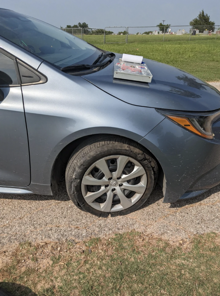 A grey car is parked on a gravel path with a flat front tire. Two automotive books are placed on the car's hood. There is green grass in the background and a chain-link fence enclosing an open field. The image is taken in a rural or suburban area.