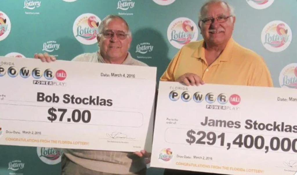 Two men stand side by side holding oversized Florida Lottery checks. The man on the left, Bob Stocklas, holds a check for $7.00 dated March 4, 2016. The man on the right, James Stocklas, holds a check for $291,400,000.00 dated March 3, 2016.