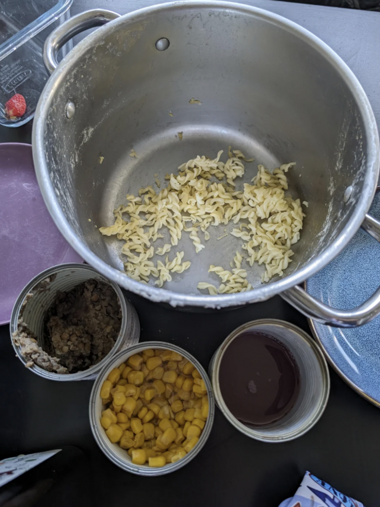 A large empty pot with a small amount of cooked pasta at the bottom is on a table. Surrounding it are three opened cans: one with ground meat, one with corn kernels, and one with a dark liquid. A blue plate sits nearby.