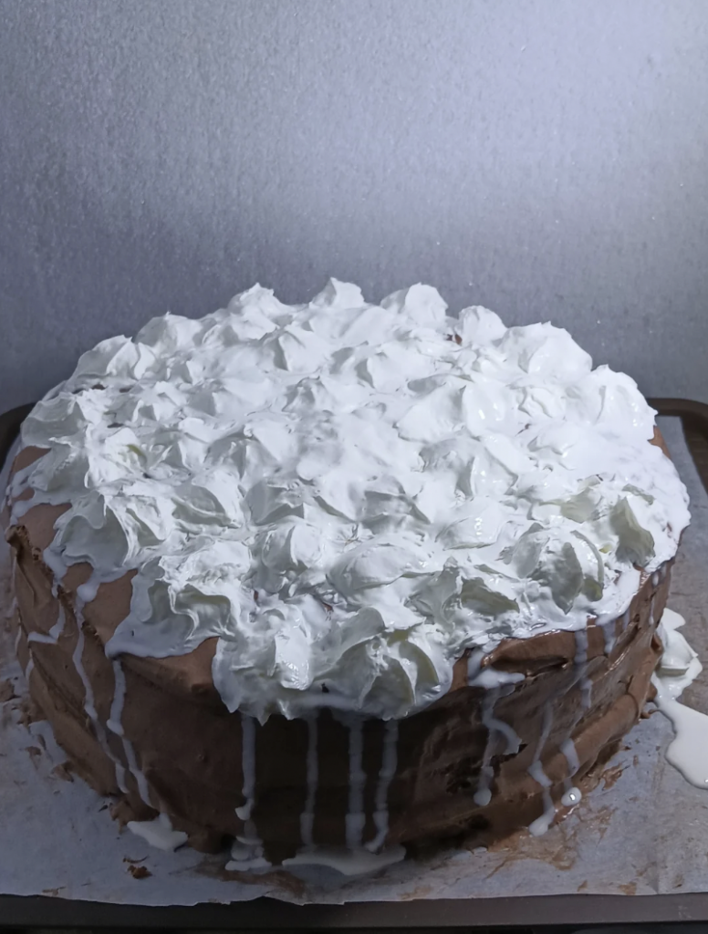 A round, homemade cake with a layer of thick white icing on top, which appears to be lumpy and uneven. Some of the icing is dripping down the sides of the cake, which is placed on a baking sheet lined with parchment paper. The background is plain and speckled.