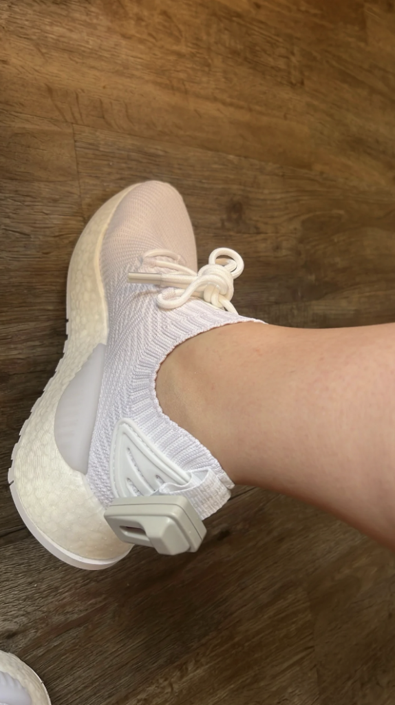 A person wearing a white sneaker with a textured pattern on a wooden floor. The shoe has a thick, cushioned sole and a white heel loop. The ankle area is snug, and a white strap with a clasp is visible at the back.