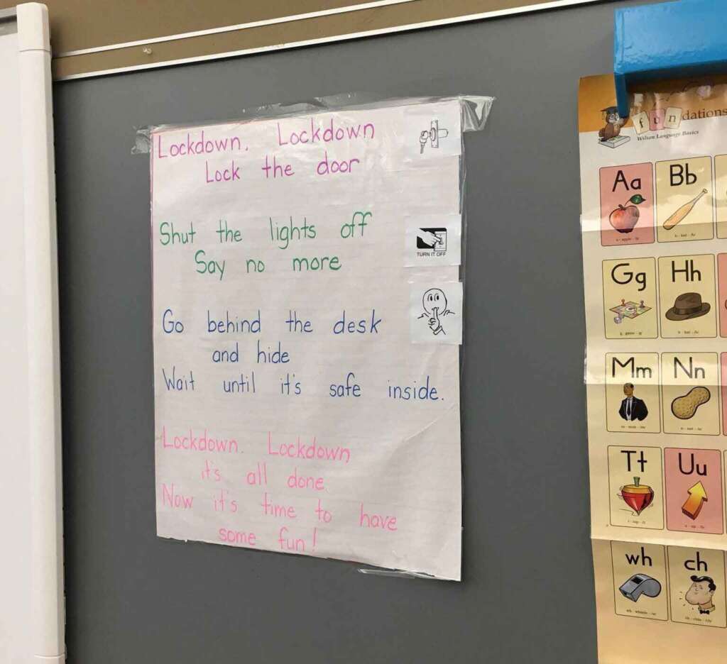 A handwritten lockdown drill poster taped to a classroom wall lists steps in four lines: "Lockdown, Lockdown, Lock the door / Shut the lights off, Say no more / Go behind the desk, Wait until it's safe inside / Lockdown, Lockdown, it's all done, Now it's time to have some fun!