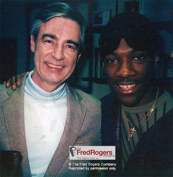 Two men are smiling and posing for a photo together. The man on the left has gray hair and is wearing a light blue turtleneck with a light brown jacket. The man on the right is wearing a black headband and a black jacket with a cross necklace.