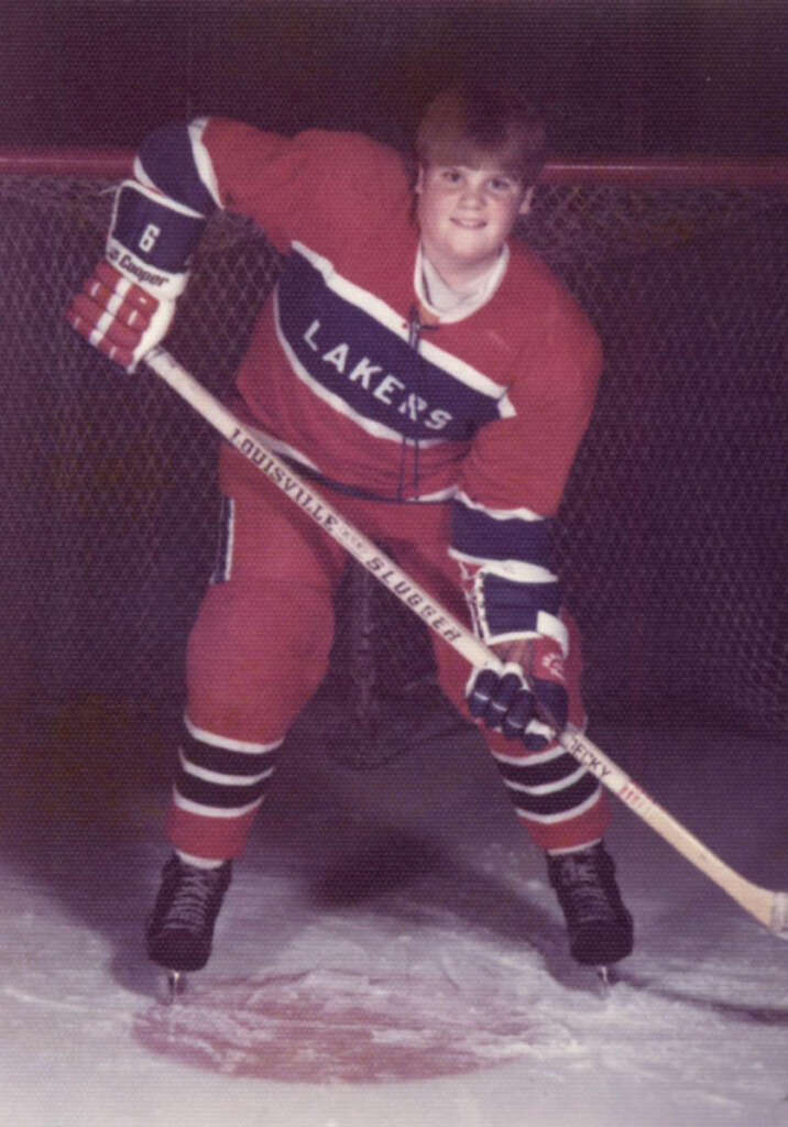 A young hockey player, wearing a red "Lakers" uniform with protective gear and skates, poses on the ice rink. He is holding a hockey stick and standing in front of a goal net, smiling at the camera.