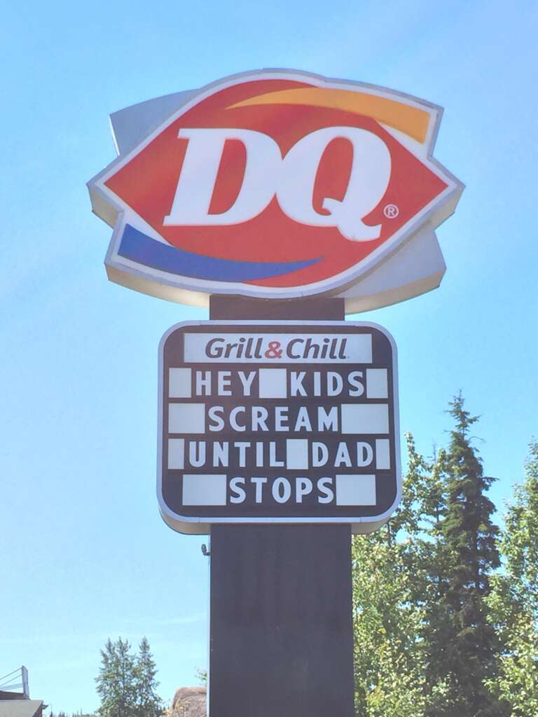 A Dairy Queen sign with the logo at the top and the words "Grill & Chill" below it. Beneath that, a marquee reads "HEY KIDS SCREAM UNTIL DAD STOPS" against a clear blue sky with some trees in the background.