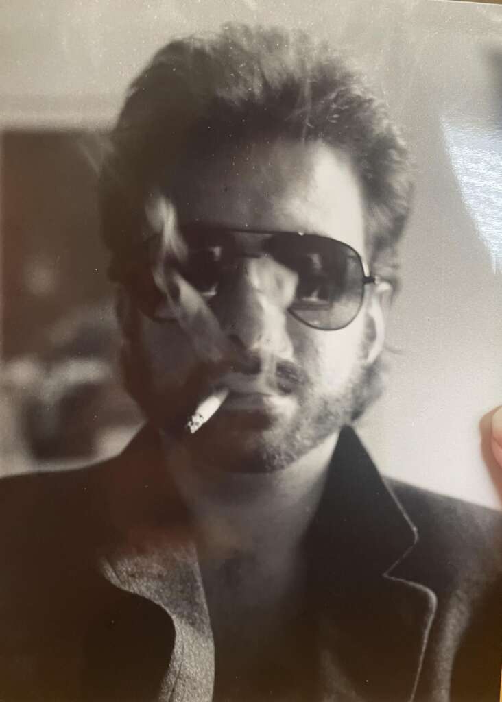 A man with thick hair and a beard wears dark sunglasses and a blazer. He is smoking a cigarette, and the smoke is visibly curling around his face. The image is in black and white, and the man's expression is serious.