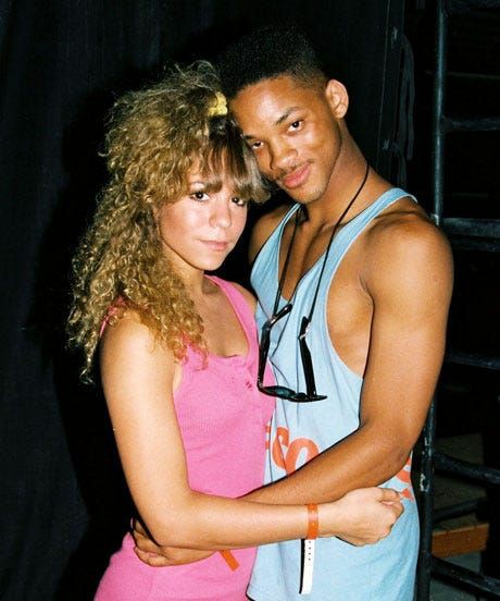 Two young adults are posing together in a warm embrace. The woman on the left has long curly hair and is wearing a pink top. The man on the right has short hair, is wearing a sleeveless blue shirt, and has sunglasses hanging from his shirt.