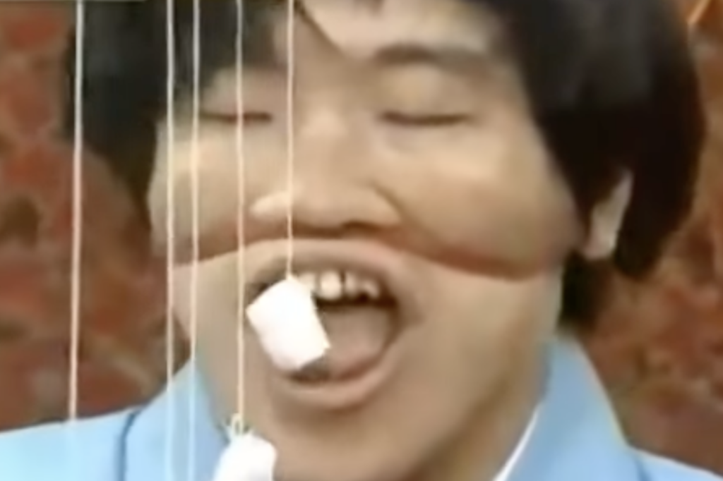 A person with closed eyes and an amused expression attempts to eat a marshmallow hanging from a string.