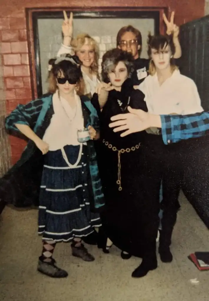 A group of five people posing indoors, dressed in various eccentric and retro outfits. The person in the center front is wearing a black dress with a gold chain belt, and others are dressed in colorful, playful clothing. Two individuals in the back are making bunny ears.