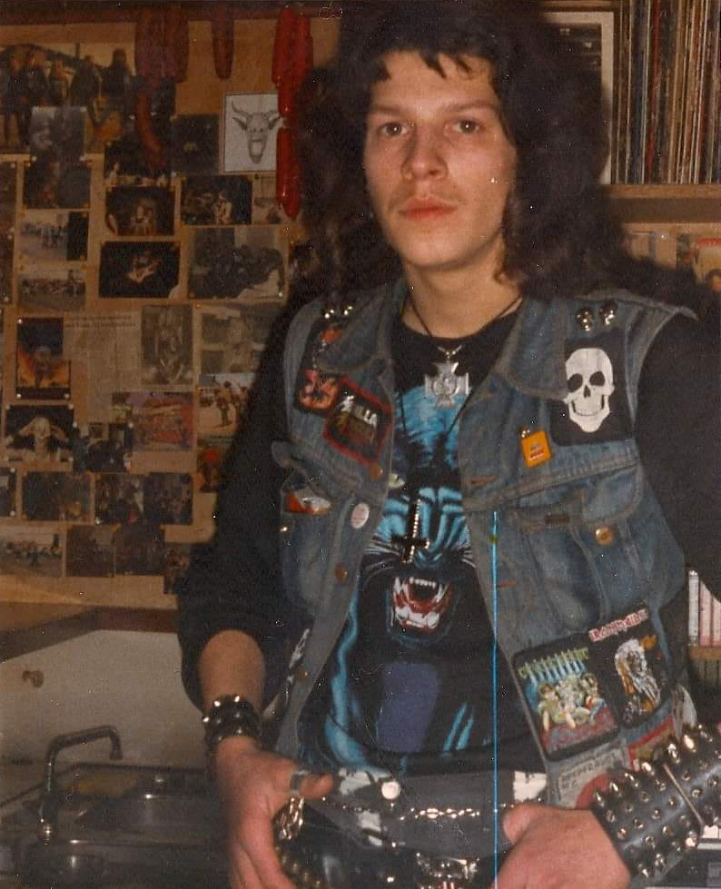 A person with long curly hair stands in a room adorned with band posters. They are wearing a patch-covered denim vest over a graphic tee, which features a snarling wolf. Their outfit includes a studded belt and bracelets, exuding a rock or punk style.