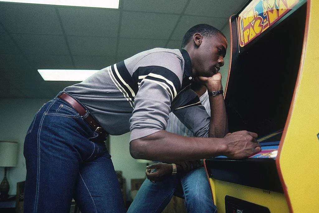 A person is intensely focused while playing an arcade game, possibly Pac-Man, in a dimly lit room. They are leaning forward with one hand on the joystick and another supporting their chin. The player's attire includes a striped shirt and blue jeans.