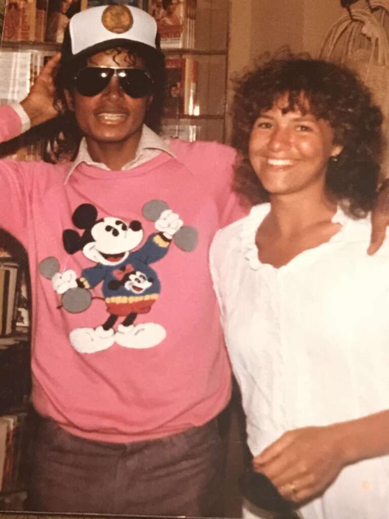 A man wearing a pink sweater with Mickey Mouse lifting weights and a white cap with a badge stands next to a smiling woman in a white blouse. The man is also wearing dark sunglasses and has his arm around the woman's shoulders. They are indoors.