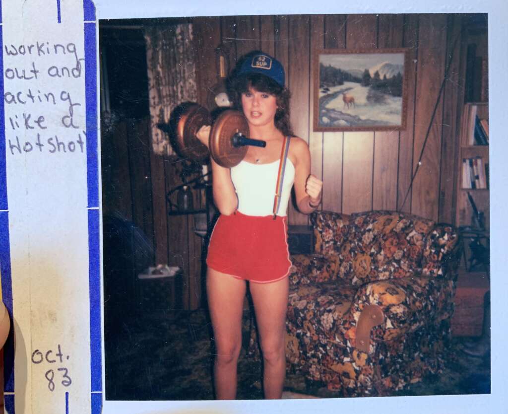 A person wearing a hat, white tank top, and red shorts is holding a dumbbell in a room with wood-paneled walls and a floral-patterned couch. A blue note on the left says, "Working out and acting like a hotshot," with "Oct. 83" at the bottom.