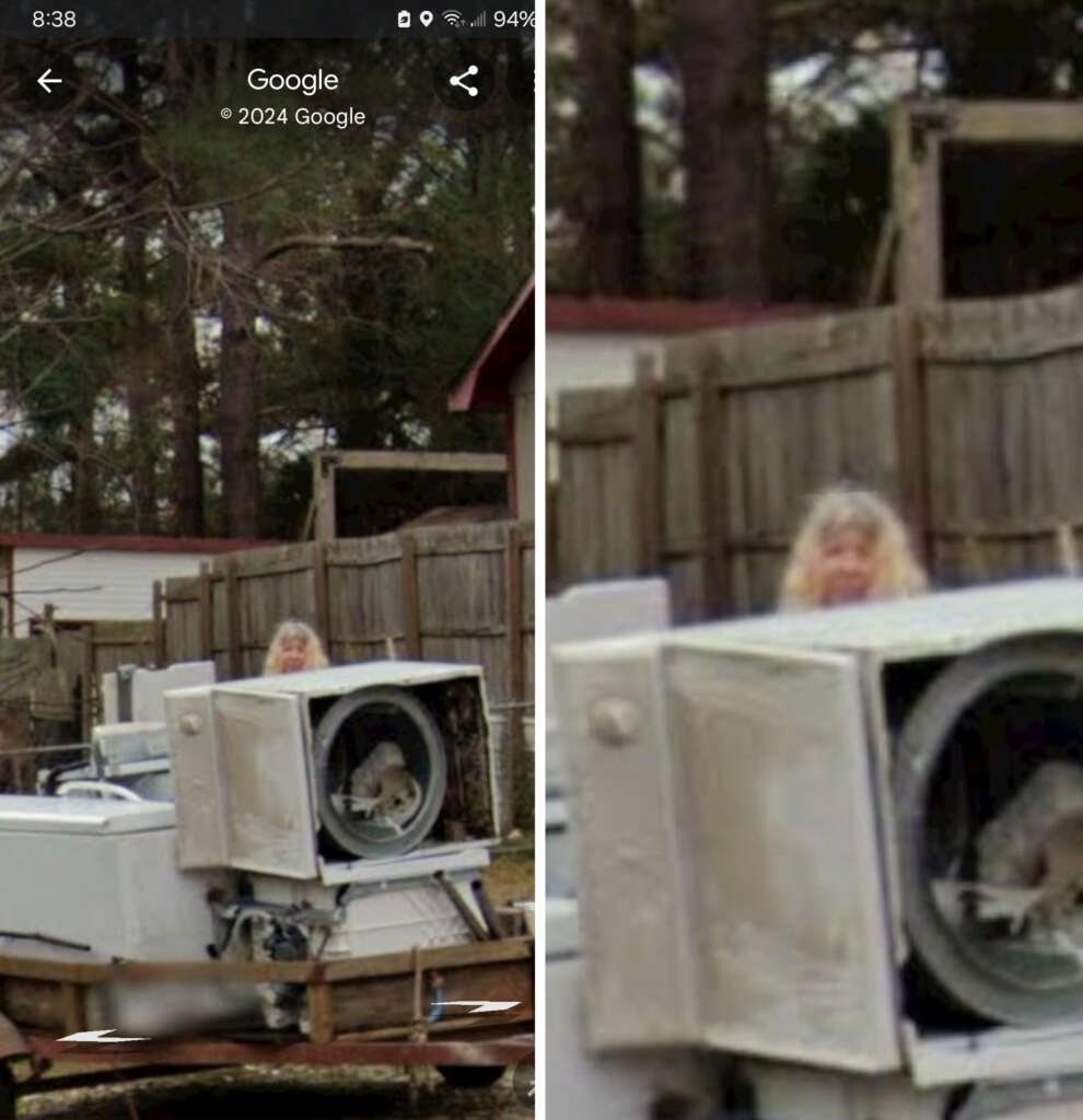 A person with long blonde hair stands outdoors beside a trailer loaded with old appliances, including a damaged washing machine. They are in a fenced area with trees and other structures visible in the background.