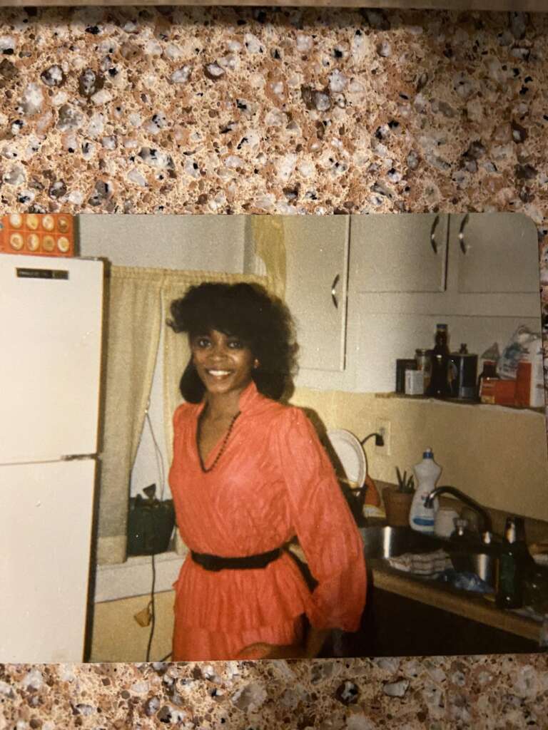 A woman with dark, curly hair, dressed in a red outfit, stands in a kitchen with a smile. The kitchen has a white refrigerator, beige countertops, cabinets, and a sink filled with dishes. Bottles and a dish soap bottle are visible near the sink.