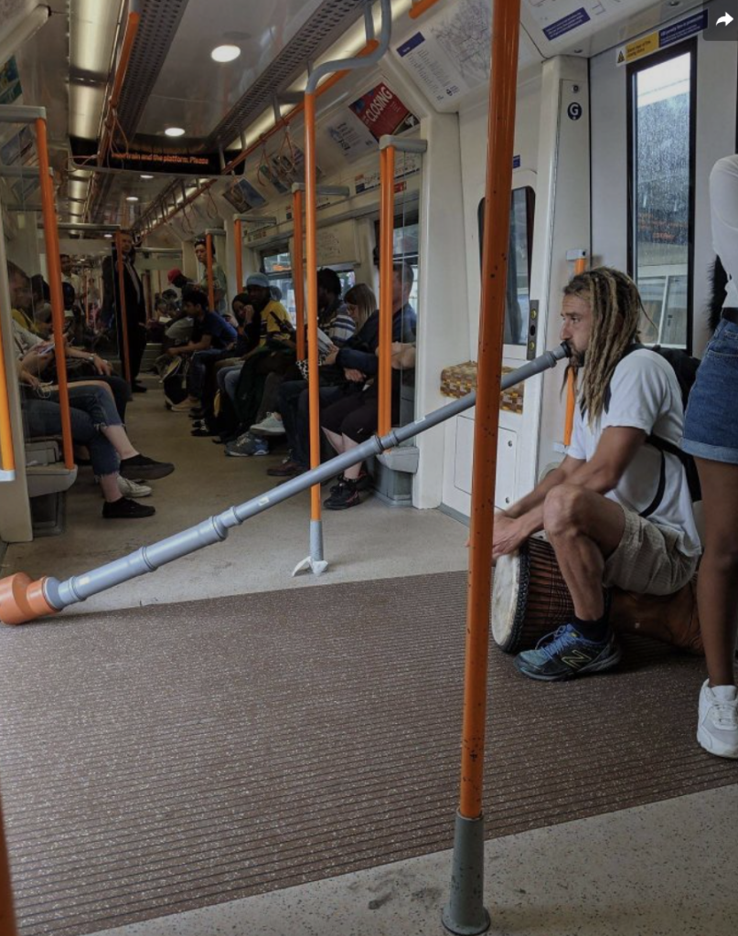 A man with long hair sits on a subway train playing a large didgeridoo and a drum. Other passengers are seated or standing, some watching him while others are engaged in their activities. The train has an orange and white color scheme, with handrails and few advertisements visible.