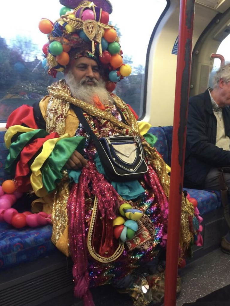 A man with a long white beard is sitting on a subway train wearing an extravagant, colorful outfit and a large, ornate headdress adorned with various decorations. The outfit is adorned with vibrant fabrics, sequins, balloons, and other accessories. Another passenger sits nearby.