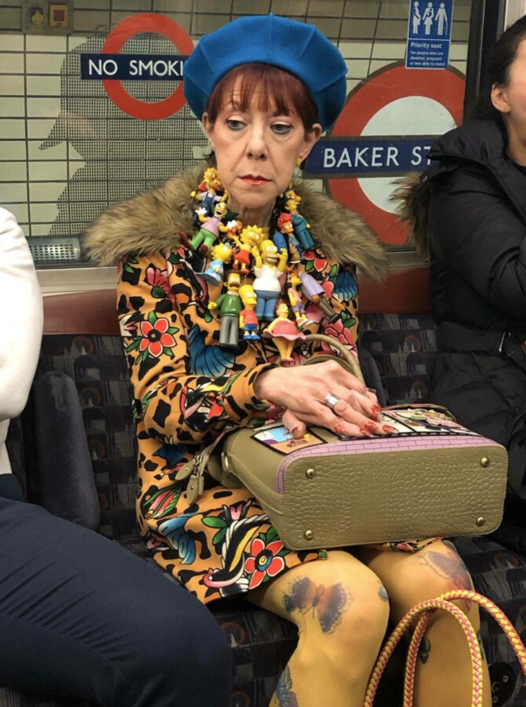 A woman sits on a subway train, wearing a blue beret, a patterned coat, and colorful tights. She has a necklace made of toy figurines and holds a beige bag on her lap. The background shows "No Smoking" and "Baker Street" signs.