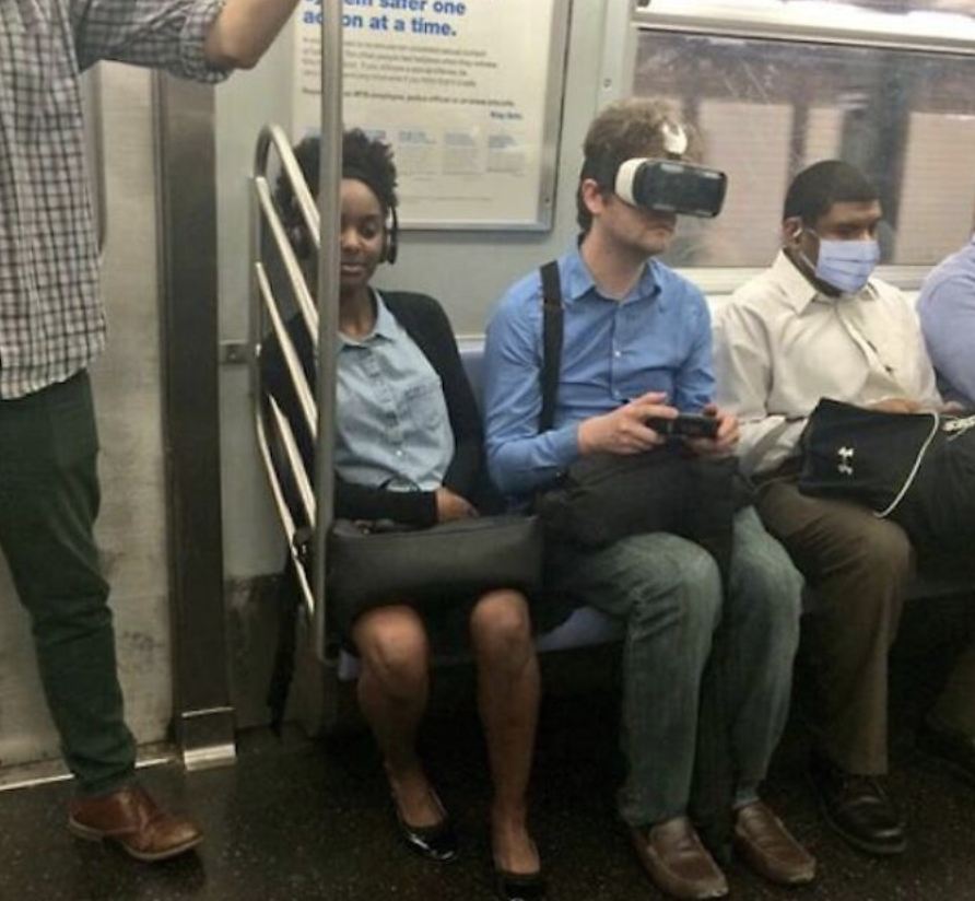 Three people sit on a subway bench. The person in the middle is wearing a VR headset and holding the controller, while the other two people are looking forward; one of them is wearing a face mask. A person standing nearby is holding onto a pole.