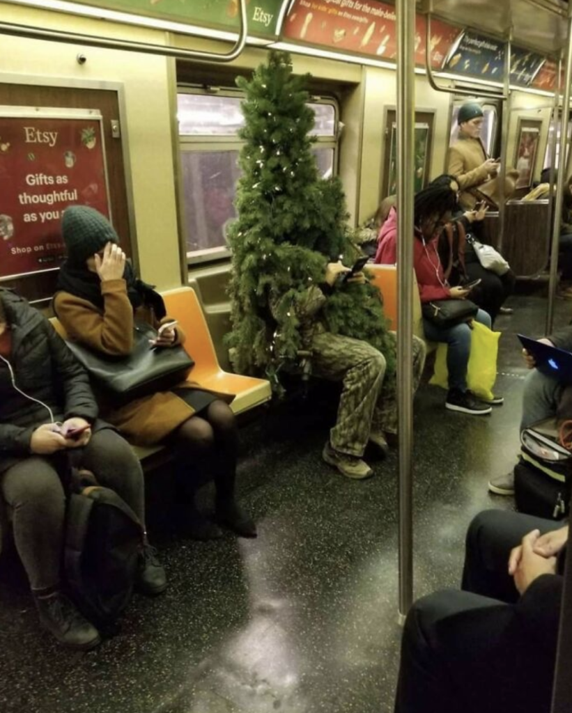 A person in a full Christmas tree costume sits on a subway seat next to other passengers. The person is barely visible through the branches. Other passengers appear calm, engaged with their own activities such as reading or using their phones.