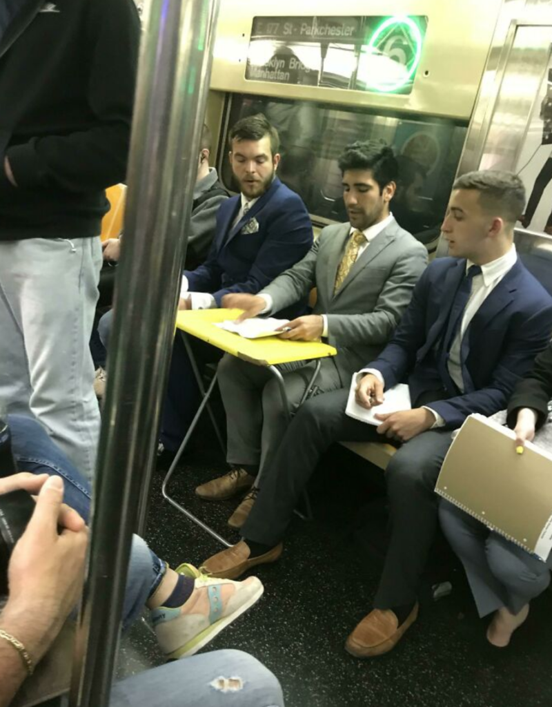 Three men in suits are seated in a subway car, engaged in conversation and looking at papers on a foldable yellow table. Other passengers are seated nearby, and subway signs are visible on the wall.