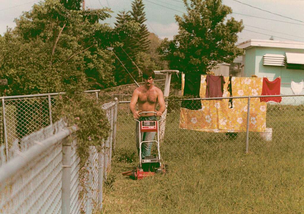 A shirtless man operates a red lawnmower in a grassy yard. Behind him, a chain-link fence separates the yard, and clothes are drying on a clothesline. Trees and a green house are visible in the background.