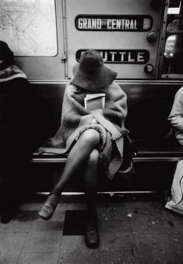 A person is sitting on a subway bench with their arms folded around a book and their legs crossed. They are wearing a hat and a poncho, partially obscuring their face. The subway sign behind them reads "Grand Central Shuttle.